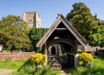 Traditional timber-framed lychgate at St Mary's Church, Salhouse, with the historic church tower visible behind and surrounded by mature trees and landscaped churchyard gardens.