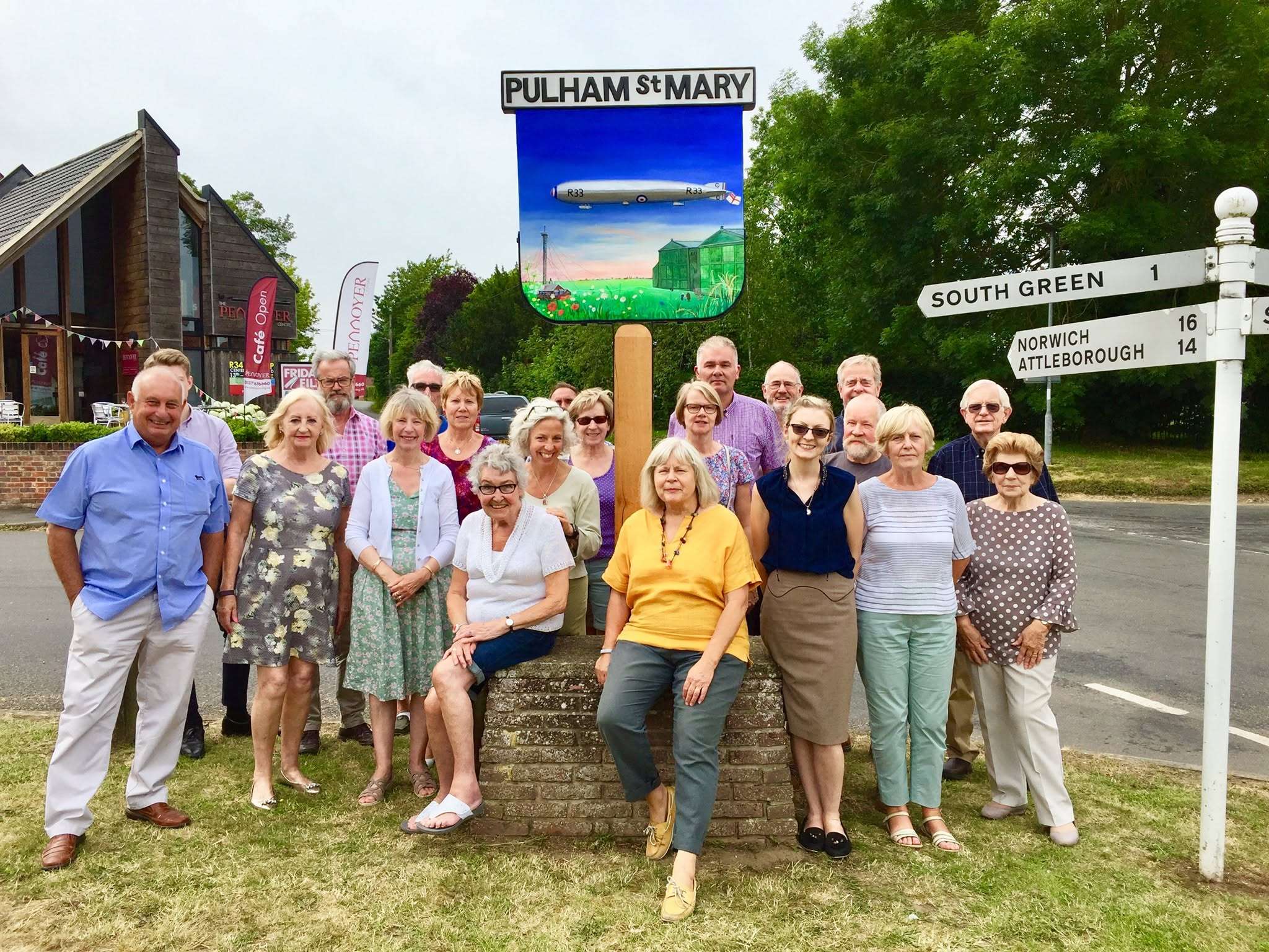 Group of people gathered beside the Pulham St Mary village sign featuring local artwork and the text “Pulham St Mary” and “South Green”.