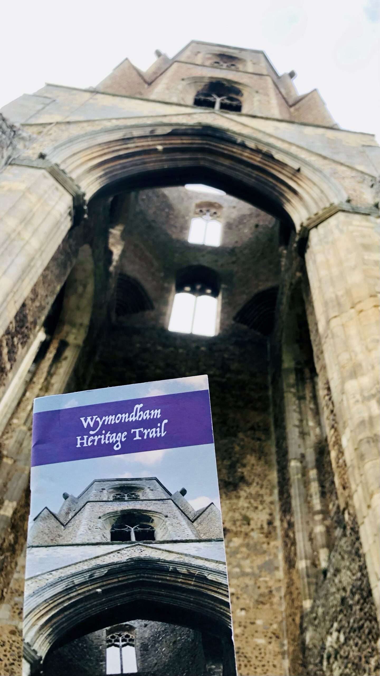 Wymondham Heritage Trail guide held inside the stone interior of Wymondham Abbey, looking up towards arched windows.