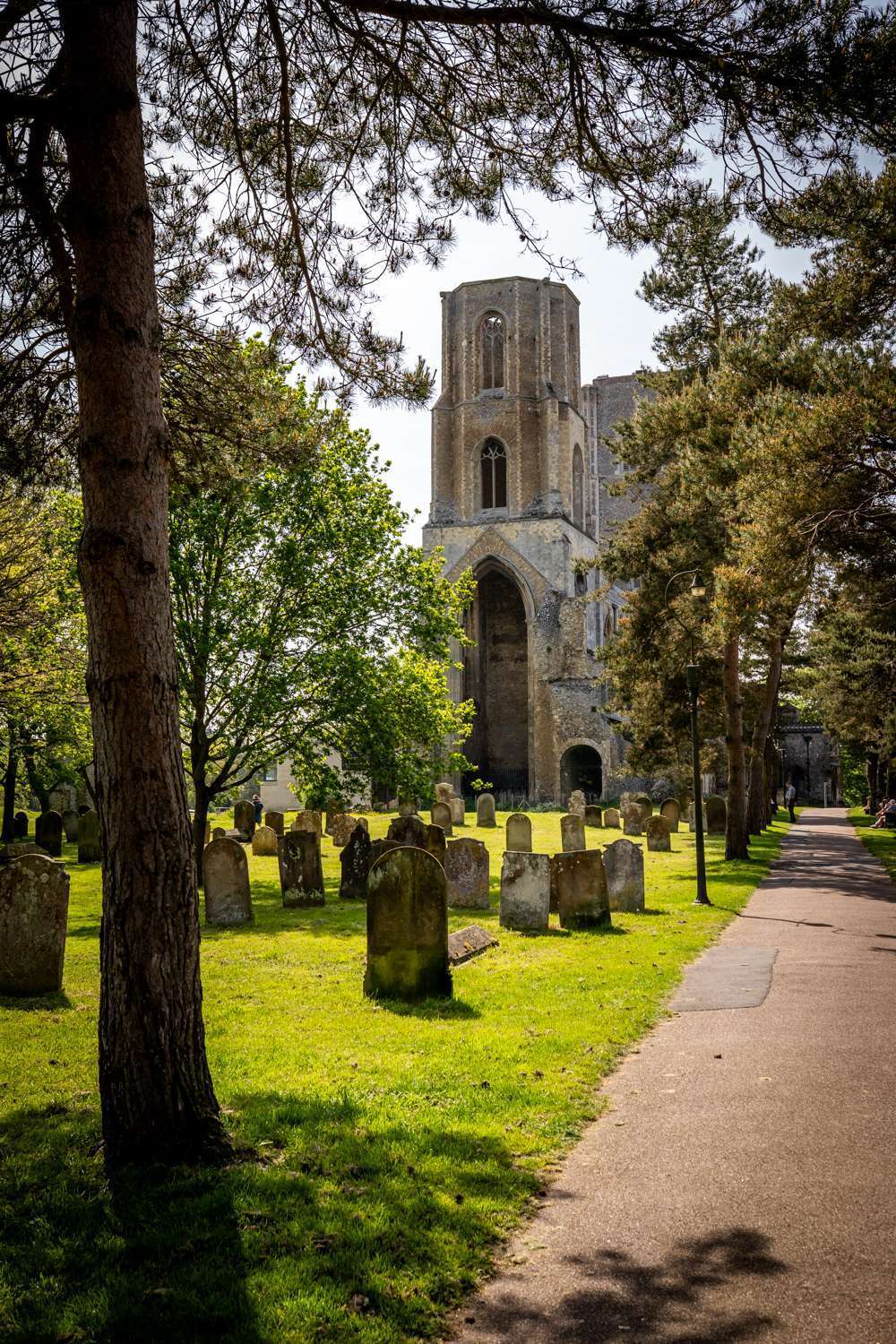 View of Wymondham Abbey tower and churchyard seen through mature trees along a pathway.