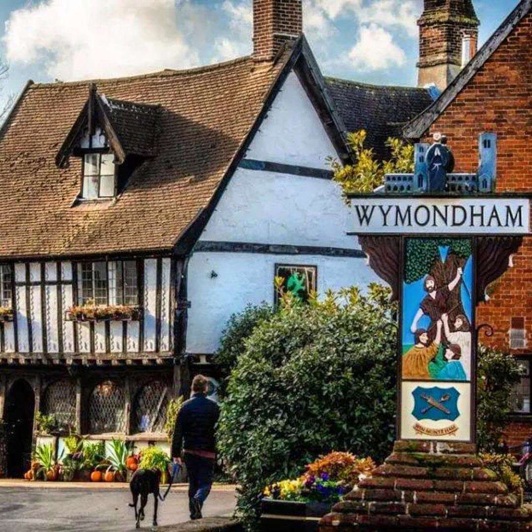 Historic timber-framed building and traditional shopfront on a street in Wymondham, with a decorative town sign displaying “Wymondham”.